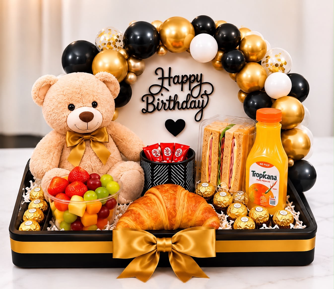 Bandeja de desayuno sorpresa con peluche, arco de globos negros y dorados, frutas, croissant y jugo